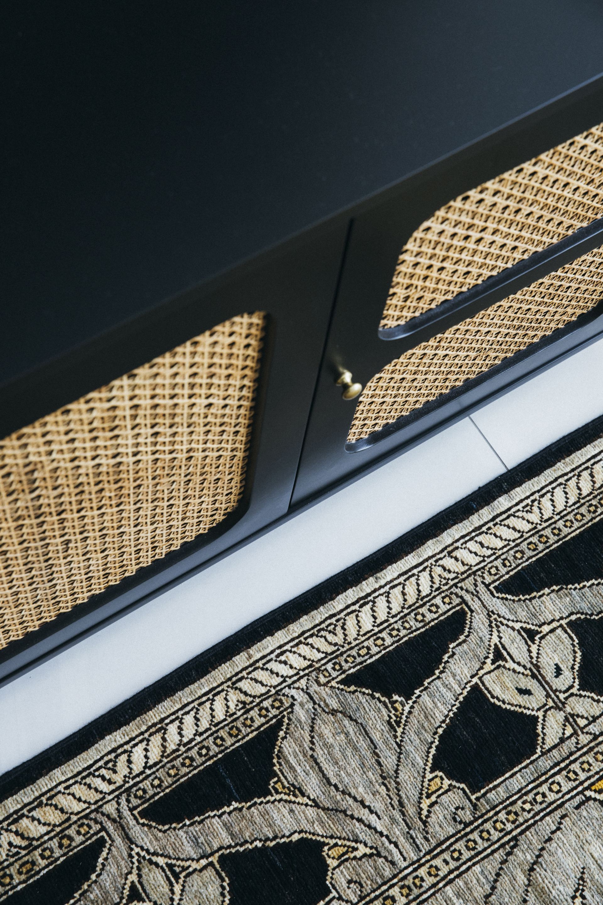 Detail of a rattan cabinet door and brass handle beside a patterned Persian rug border
