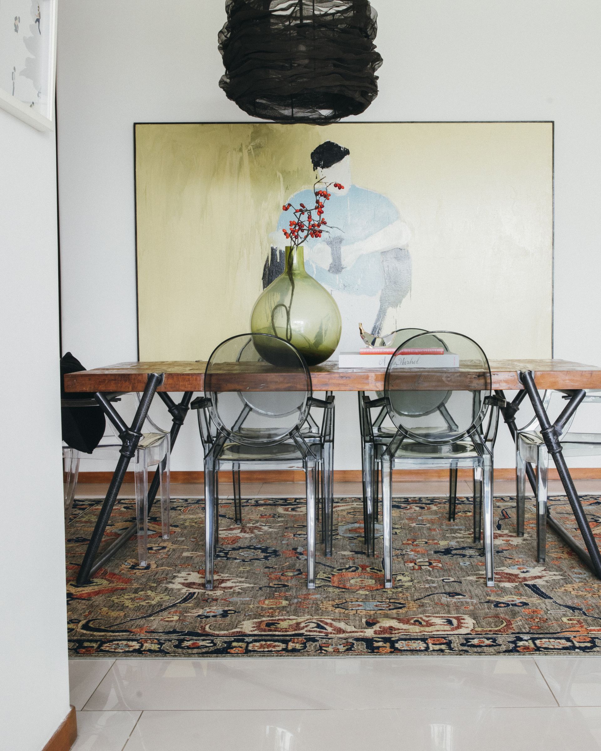 Acrylic dining chairs around a wood table beneath a large figurative painting, set on a Persian rug