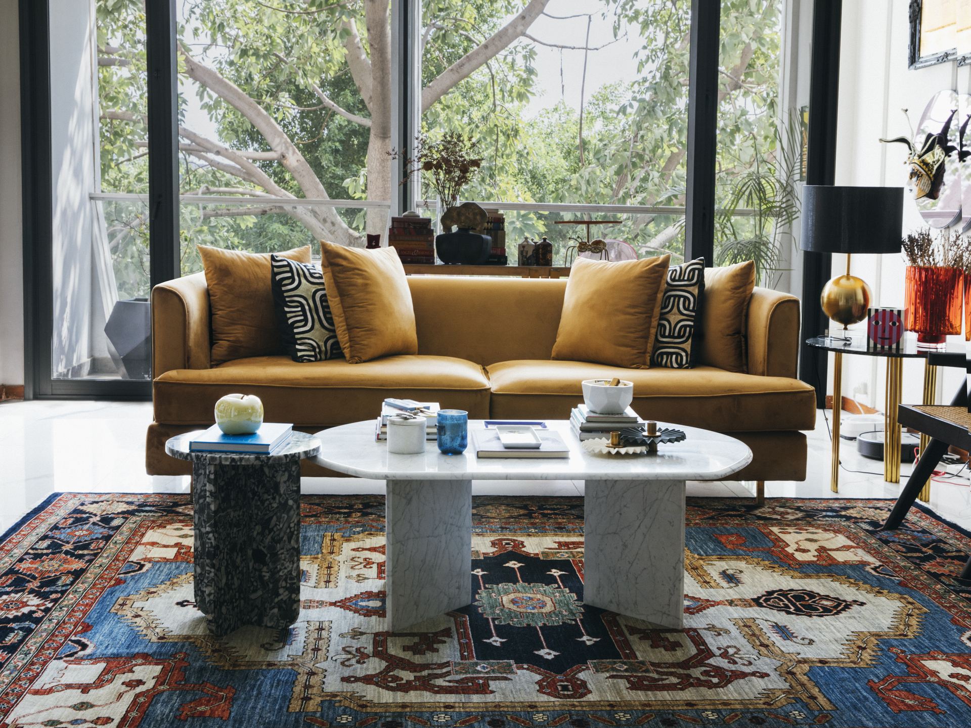 Mustard velvet sofa with graphic cushions beside marble coffee tables on a blue Persian rug