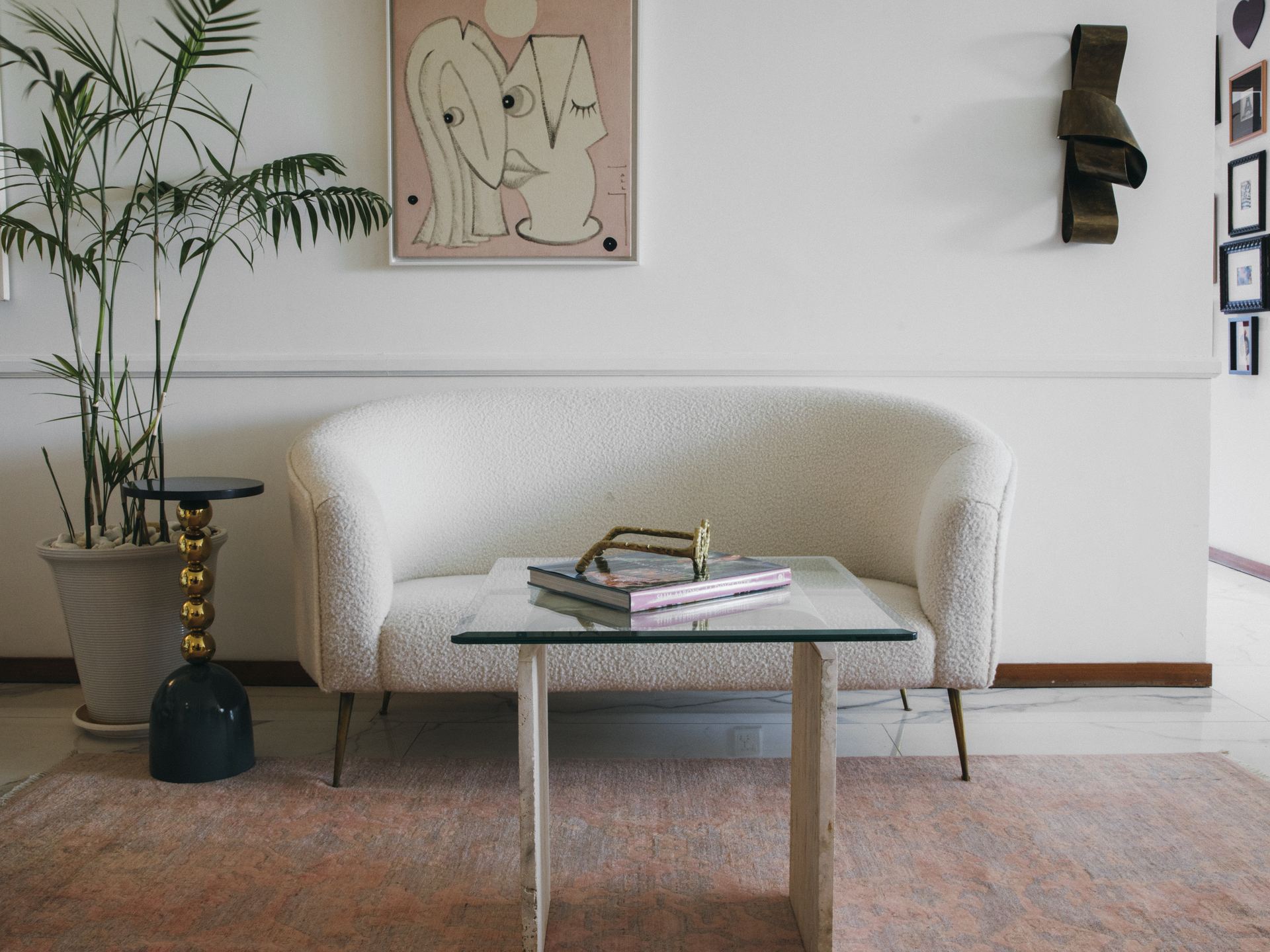 White bouclé loveseat beside a potted palm and framed illustration on a terracotta Persian rug
