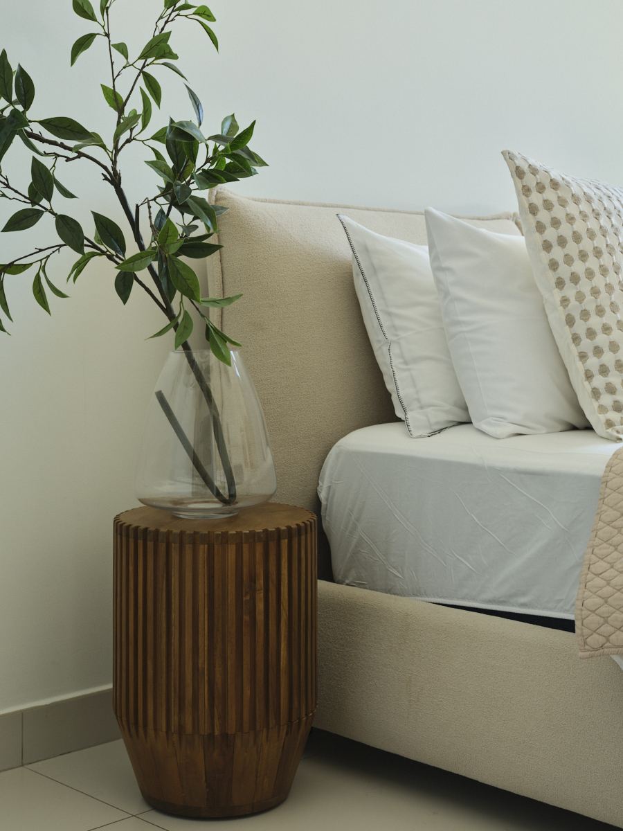 Fluted wood side table with a glass vase beside an upholstered bed