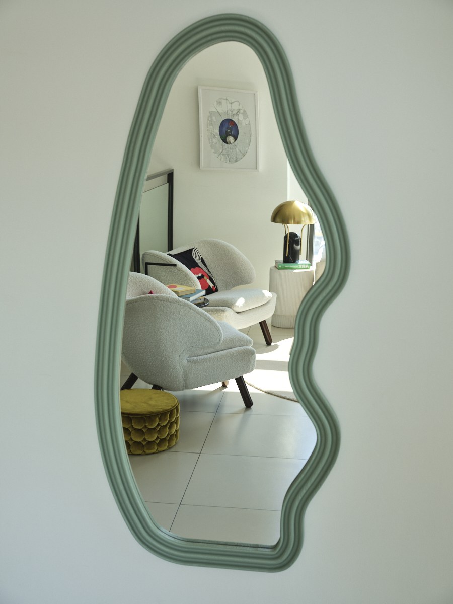 Organic-shaped sage mirror reflecting a bouclé armchair and brass table lamp