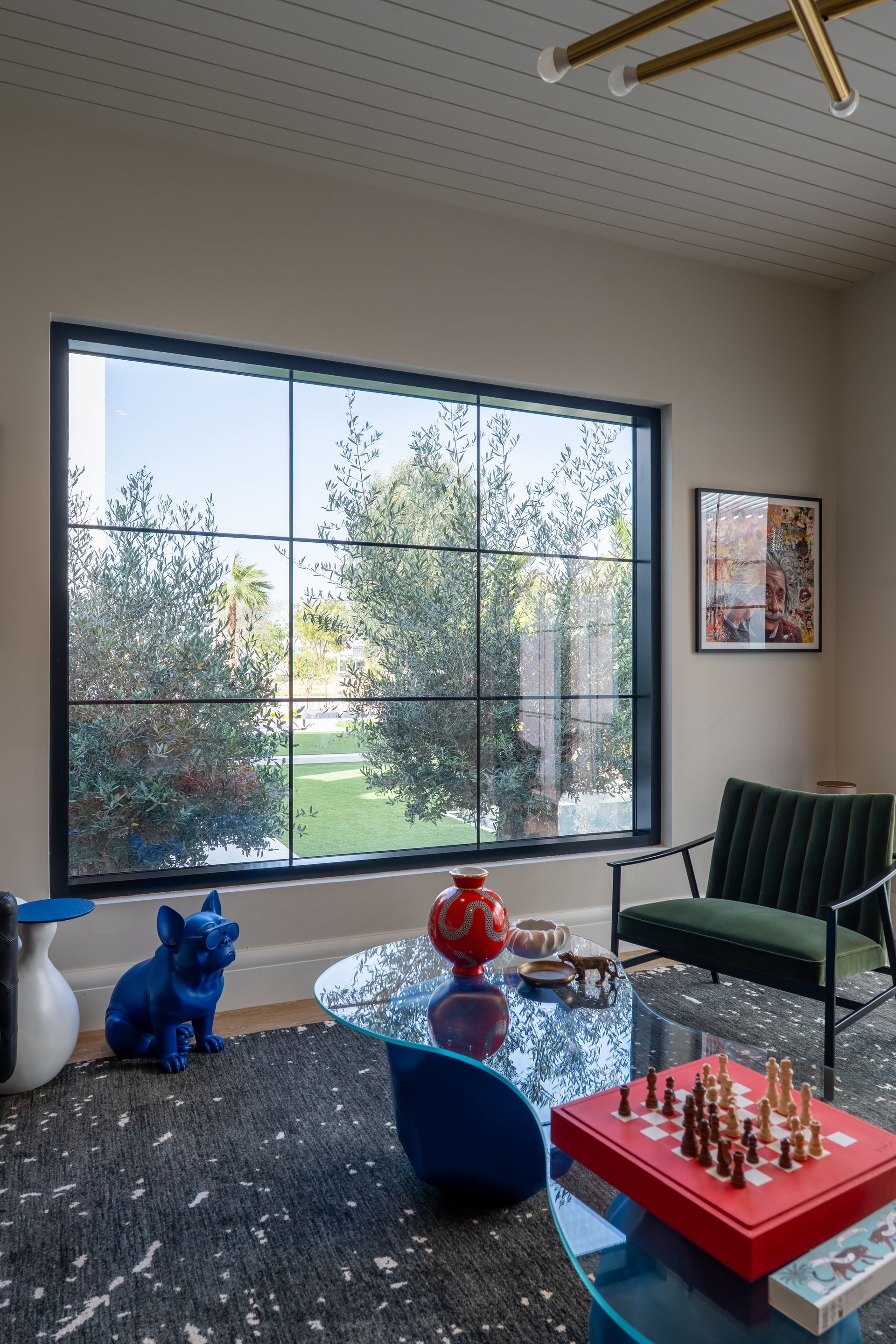Green velvet armchair beside a floor-to-ceiling window framing olive trees, with a chess set