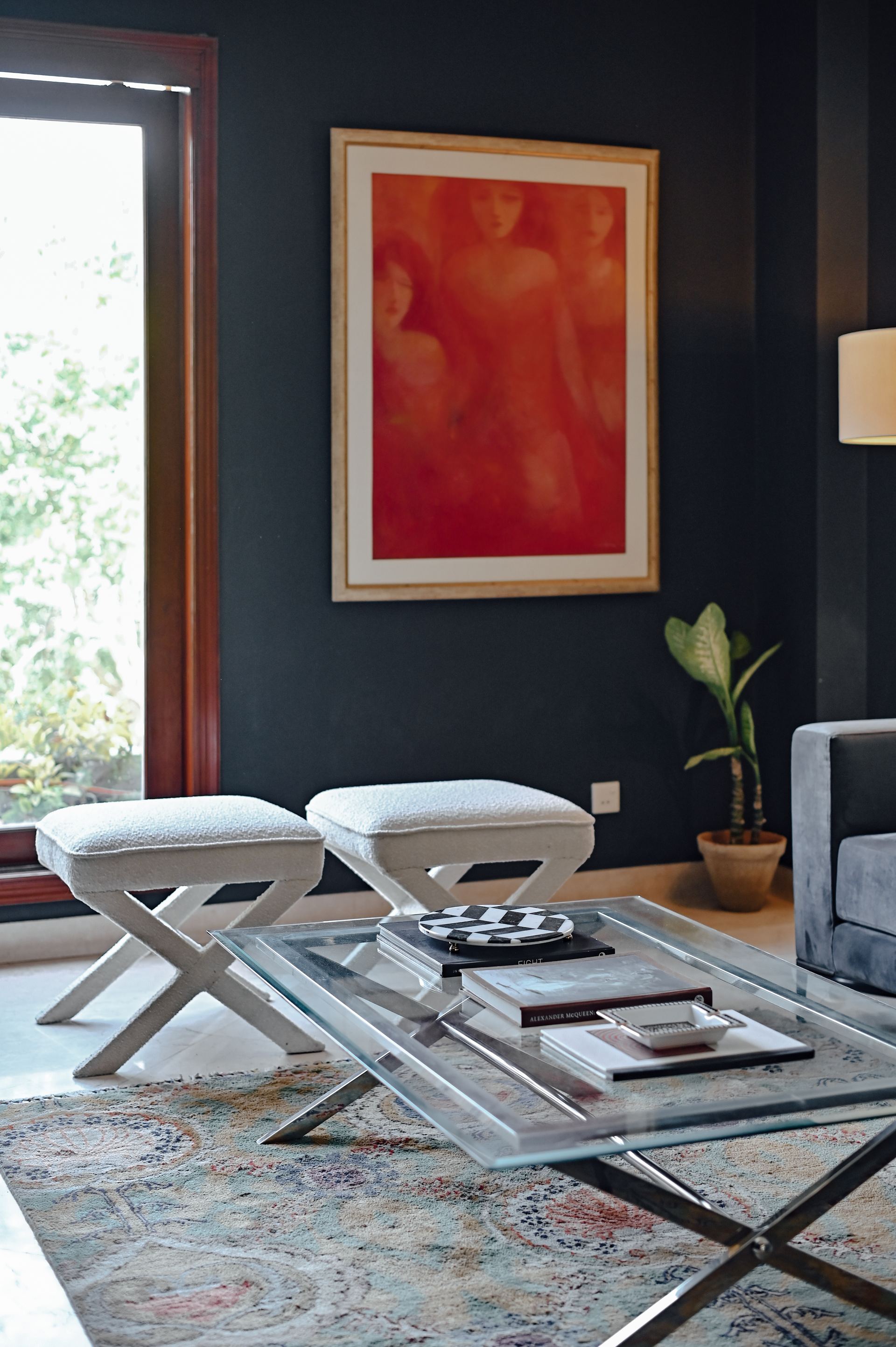 Red-toned portrait painting above cream X-frame stools and a glass coffee table