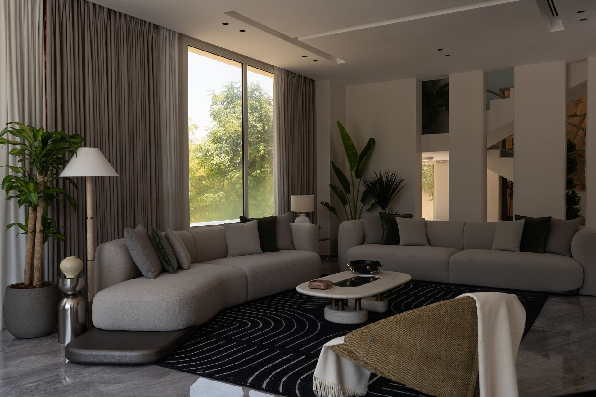 Curved grey sectional sofas around a striped rug and marble coffee table facing a garden view