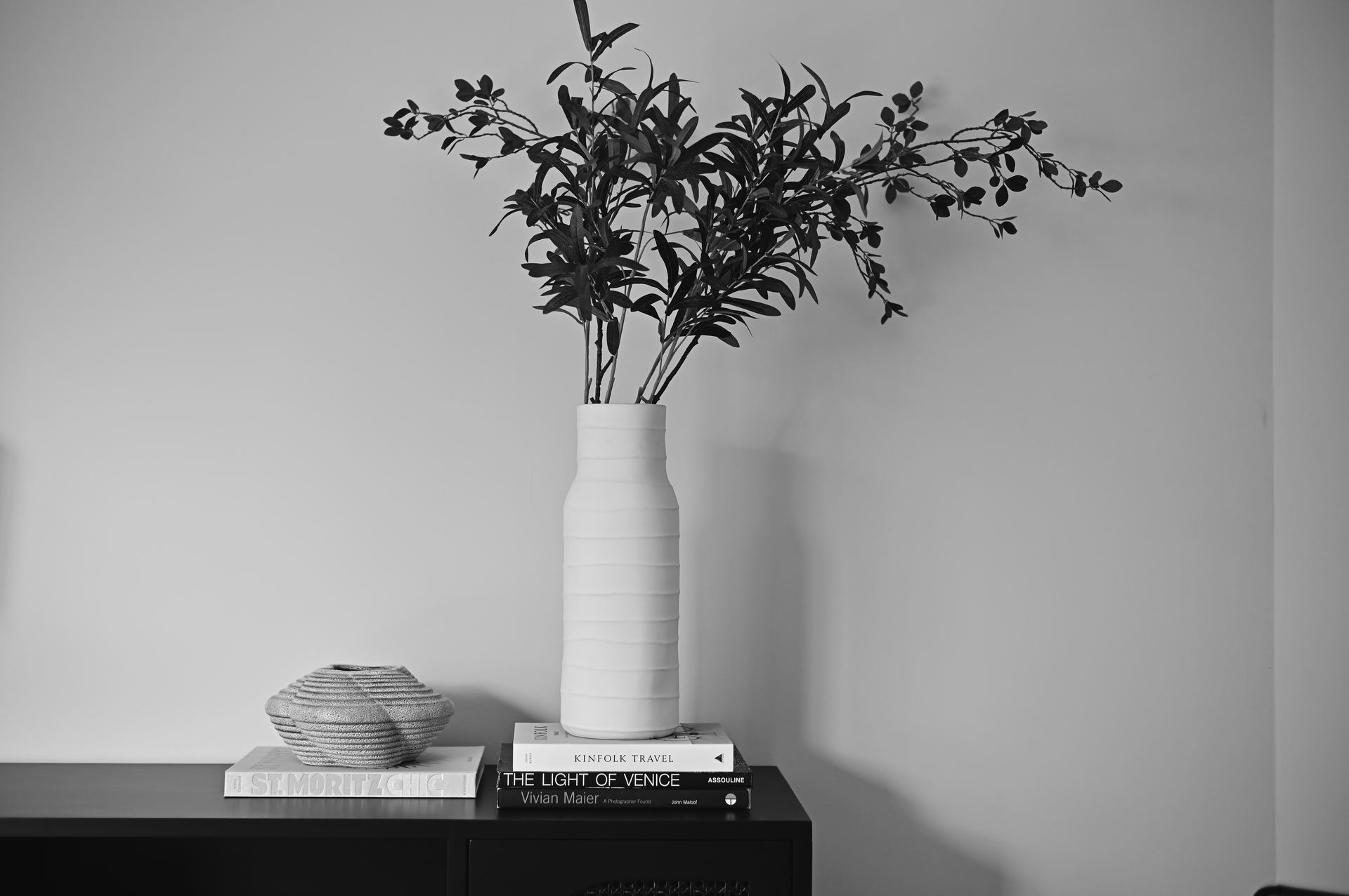 Black-and-white detail of a ceramic vase of olive branches on a black console