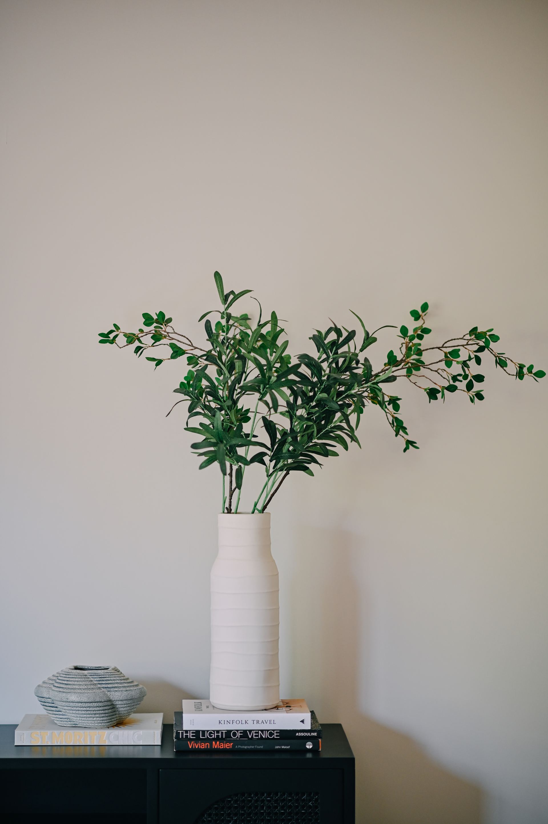 Ceramic vase of olive branches beside a sculpture and stacked books on a black console