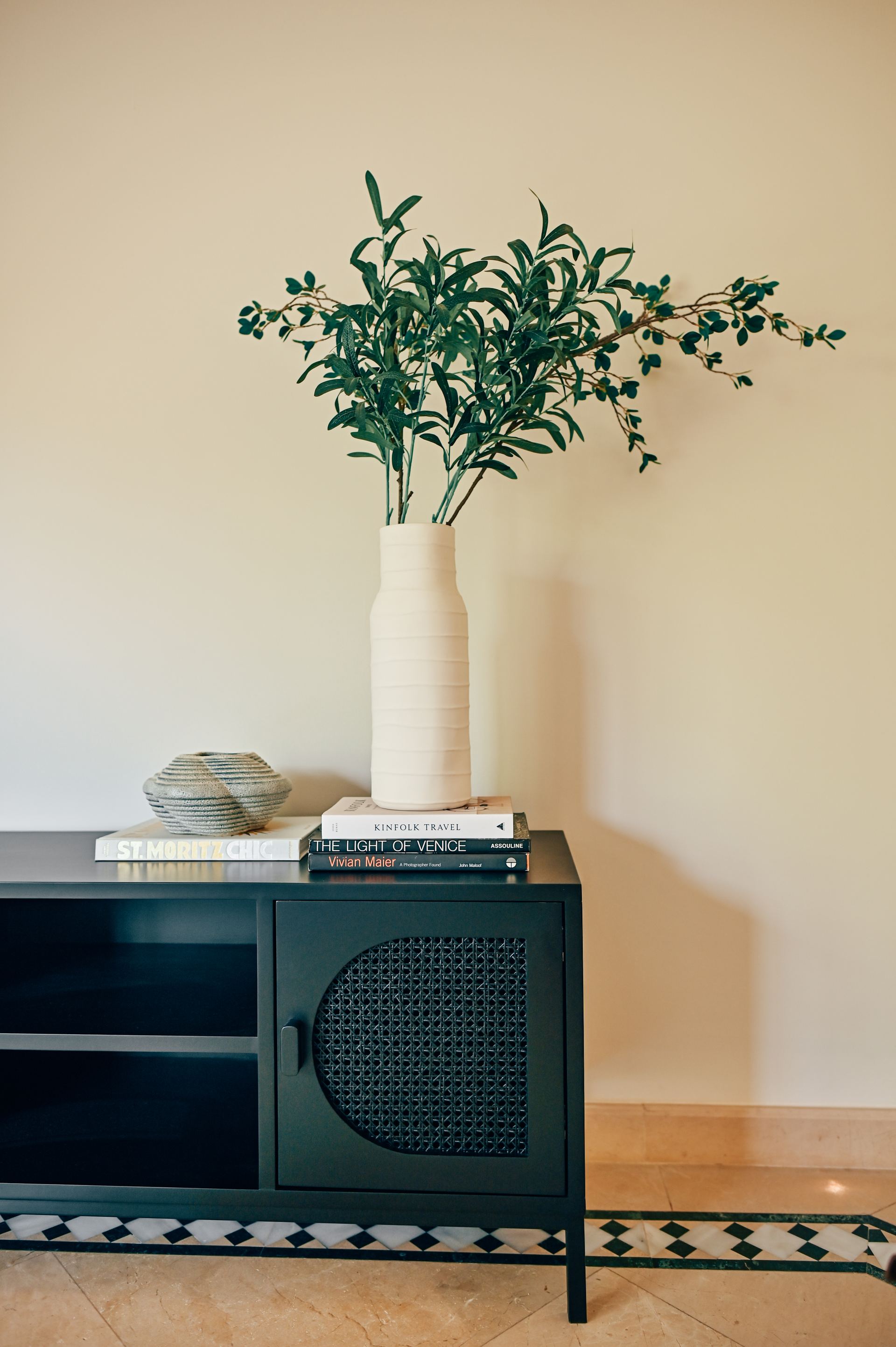 Wide view of a ceramic vase of olive branches on a black console styled with books